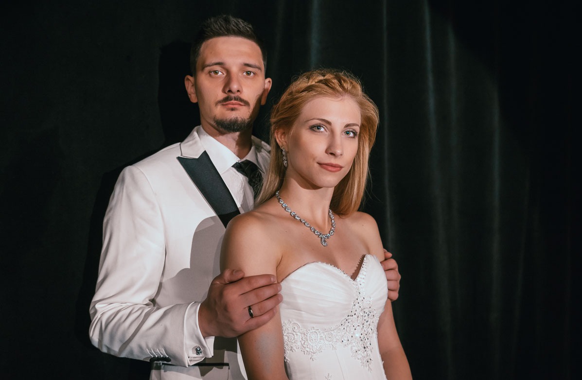 Elegancka para: blondwłosa panna młoda w białej sukni z diamentową biżuterią i pan młody w białym smokingu z czarną muchą, pozujący na tle czarnej zasłony.