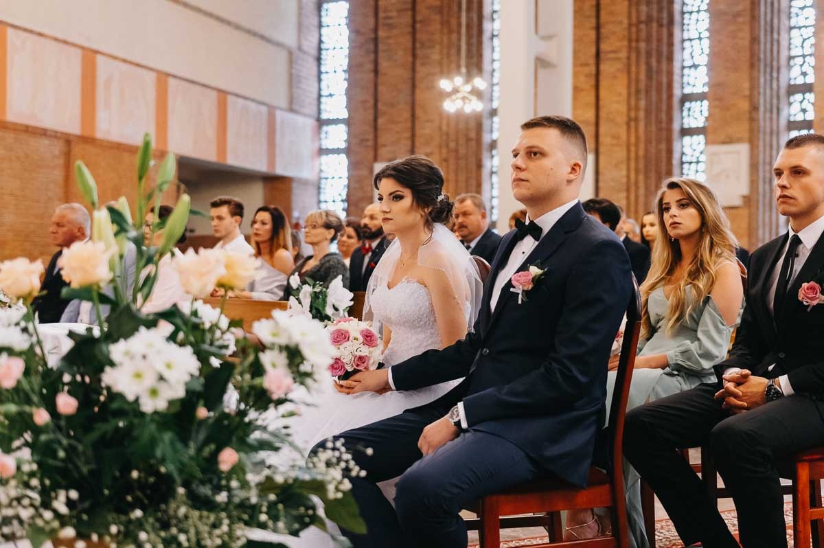 Para młoda siedzi w kościele podczas ceremonii, w tle widoczni goście, w pierwszym planie bukiet kwiatów.