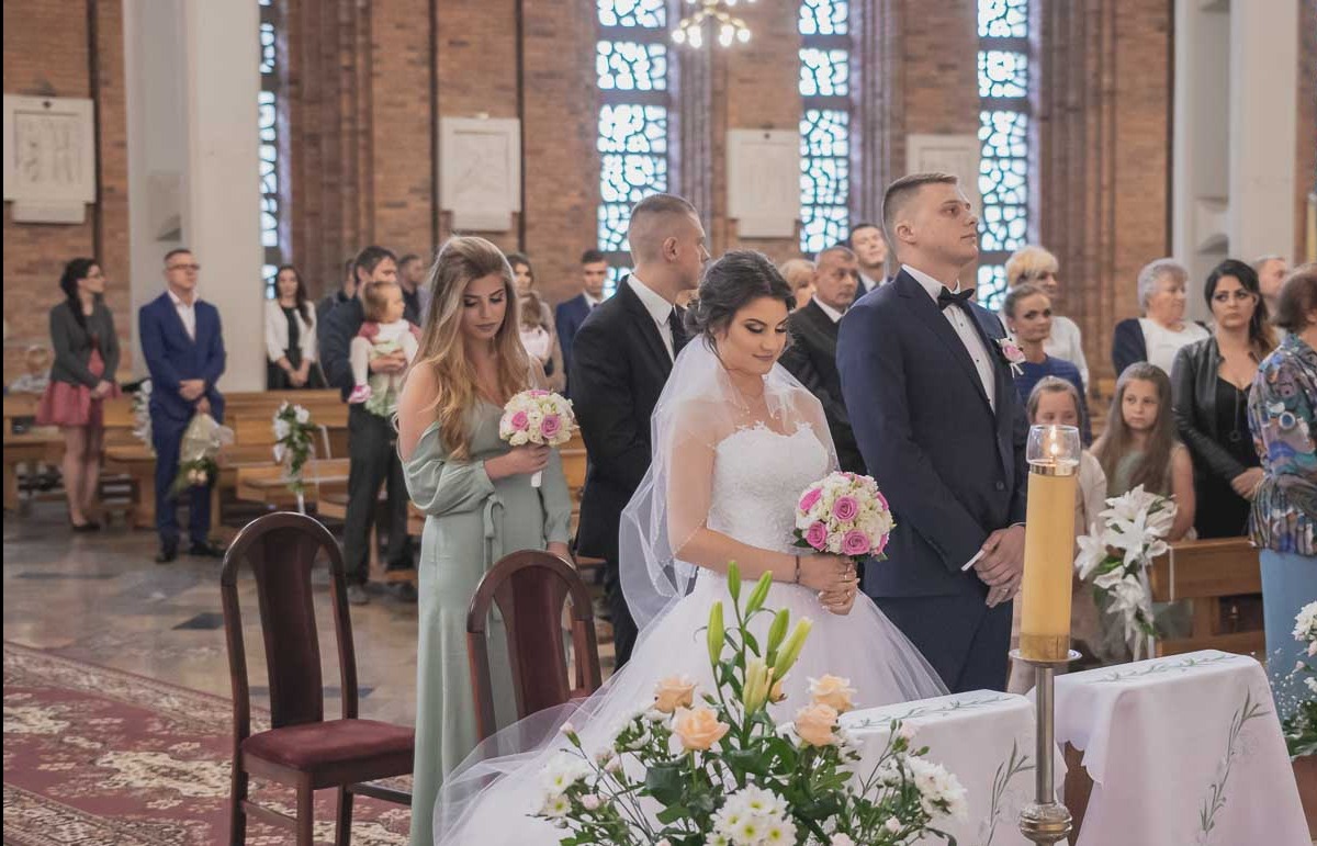 Panna młoda w białej sukni i welonie, z bukietem różowych i białych róż w dłoniach, stoi obok świadkowej w zielonej sukni, podczas ceremonii ślubnej w kościele, w tle goście.