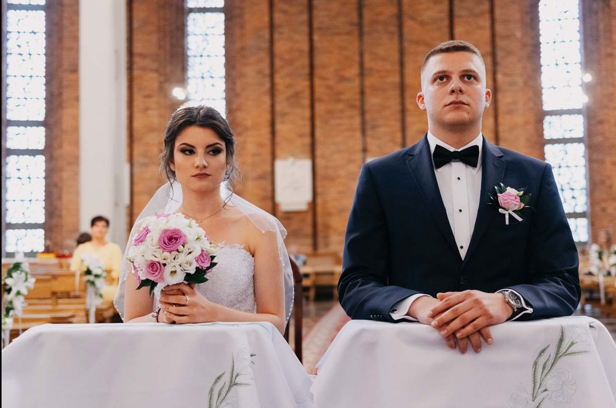 Para młoda w kościele podczas ceremonii ślubnej, panna młoda trzyma bukiet różowo-białych kwiatów, pan młody w granatowym garniturze z muchą.