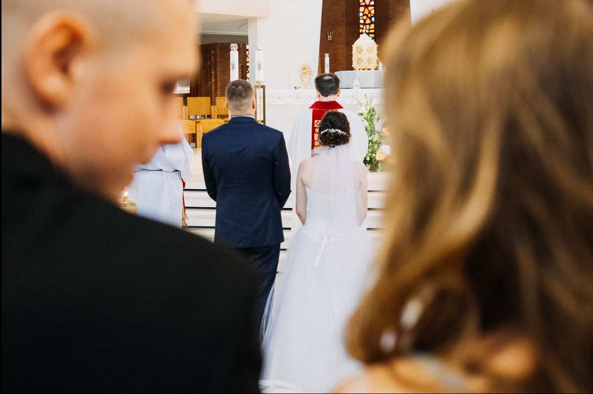 Para młoda podczas ceremonii ślubnej w kościele, widok zza ramienia gościa, skupienie na sukni panny młodej i plecach pana młodego.