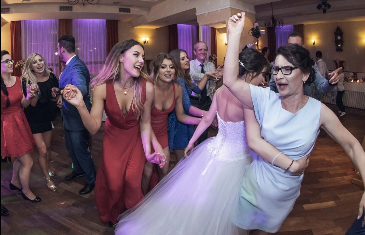 Roztańczona panna młoda w białej sukni z druhnami w czerwonych i niebieskich sukienkach oraz gośćmi weselnymi na parkiecie sali bankietowej z drewnianą podłogą i fioletowym oświetleniem.