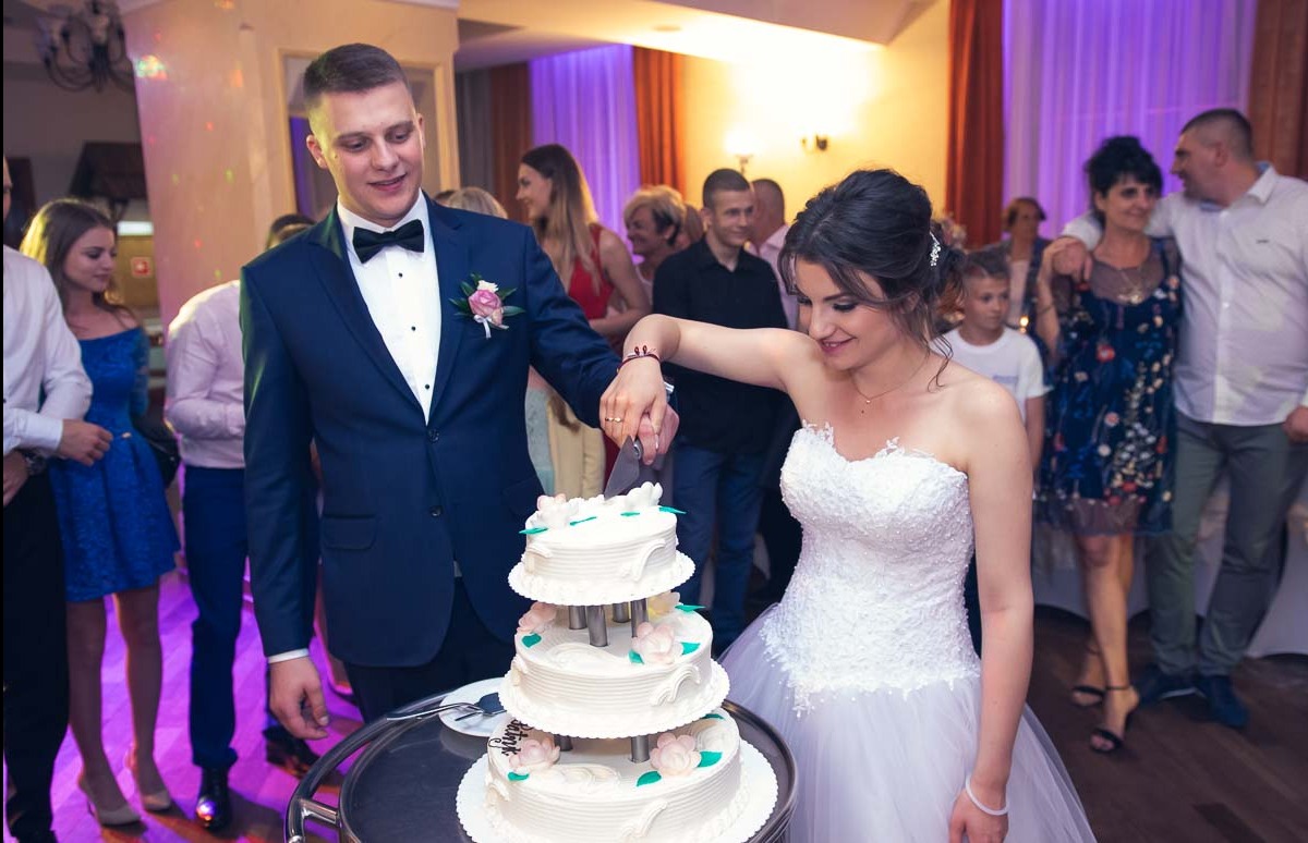 Para młoda kroi tort weselny na tle gości, ona w białej sukni bez ramiączek, on w granatowym garniturze z muchą i butonierką z różowym kwiatem.