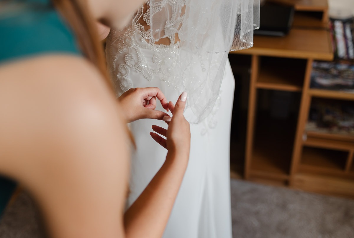 Pomoc druhny przy zapinaniu delikatnej koronki na sukni ślubnej panny młodej, widoczny fragment welonu i drewniany regał w tle.