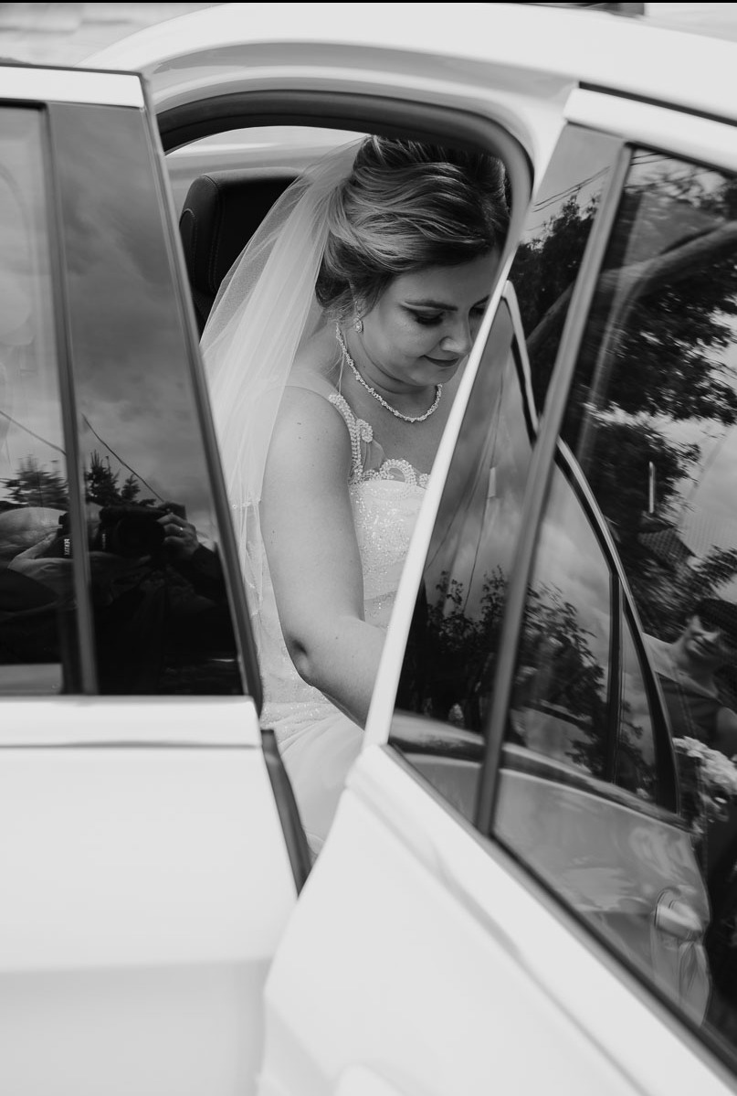 Czarno-białe zdjęcie panny młodej w sukni ślubnej wsiadającej do białego samochodu, widok przez otwarte drzwi auta.