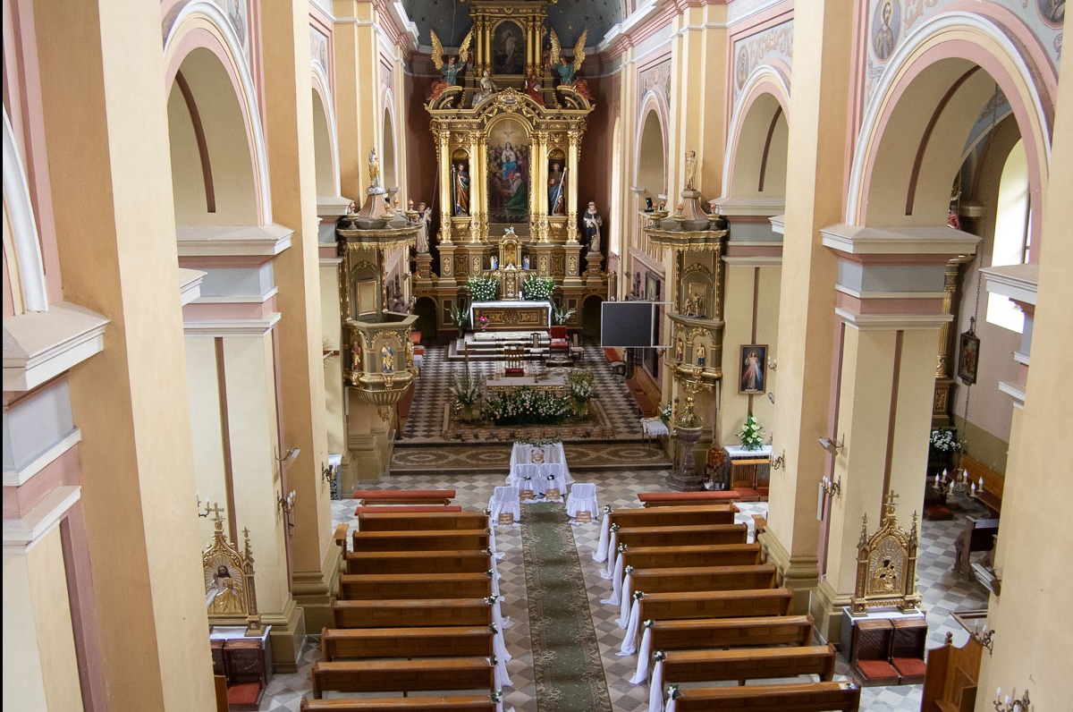 Wnętrze kościoła przygotowane do uroczystości ślubnej, widok z góry na nawę główną z dekoracjami kwiatowymi i białymi wstążkami na ławkach.
