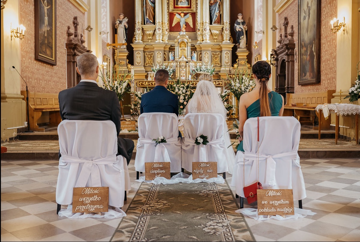 Para młoda siedząca w kościele podczas ceremonii ślubnej, widok z tyłu, krzesła udekorowane białymi pokrowcami i drewnianymi tabliczkami z cytatami o miłości.