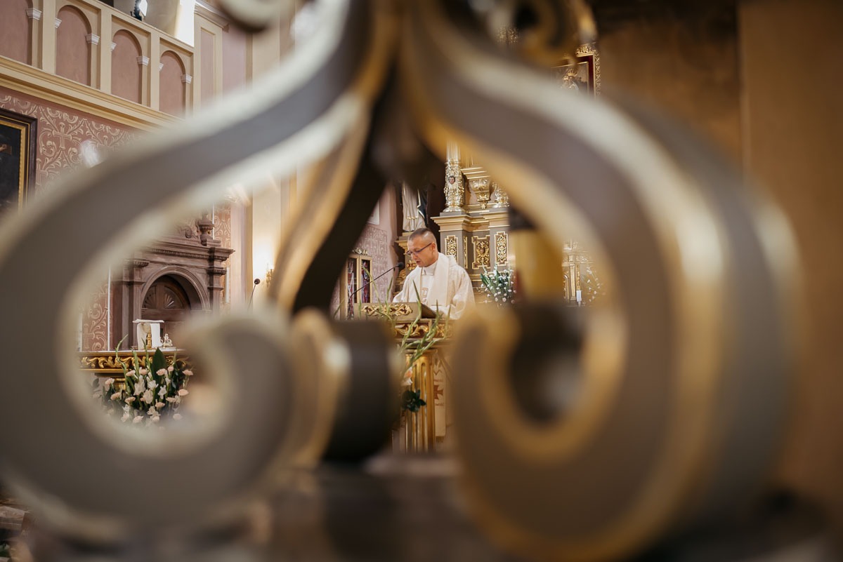 Ksiądz w ornacie czyta z pulpitu w kościele, widok przez ozdobny element balustrady.