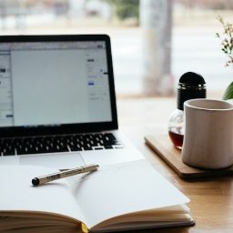 Aleksandra Smmogorzewska - Otwarty notatnik z piórem na biurku obok laptopa, filiżanki i kwiatów w słoiku, w tle okno z rozmytym widokiem.