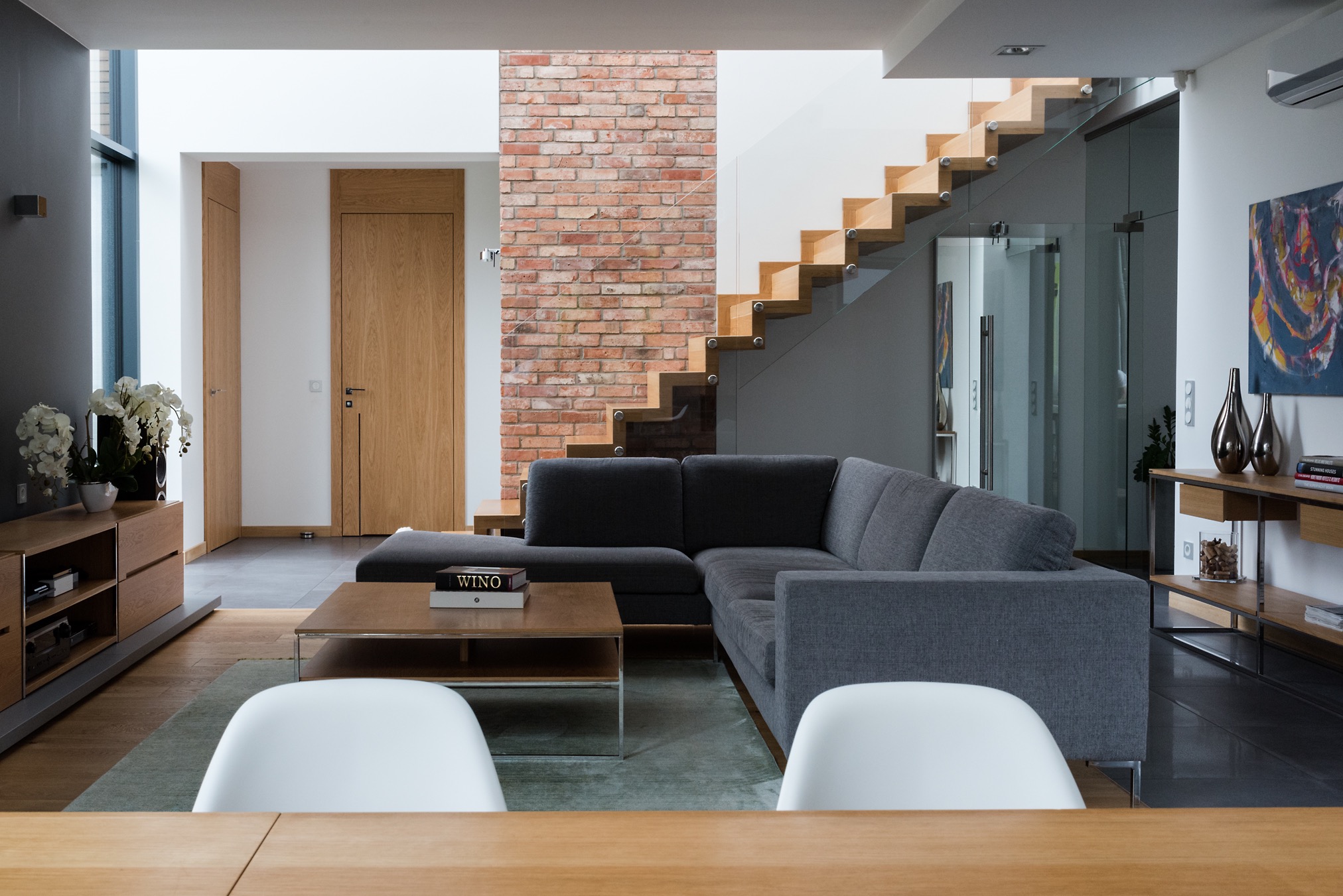 Nowoczesny salon z szarą sofą narożną, drewnianymi schodami z szklaną balustradą, ścianą z czerwonej cegły i minimalistycznymi meblami.