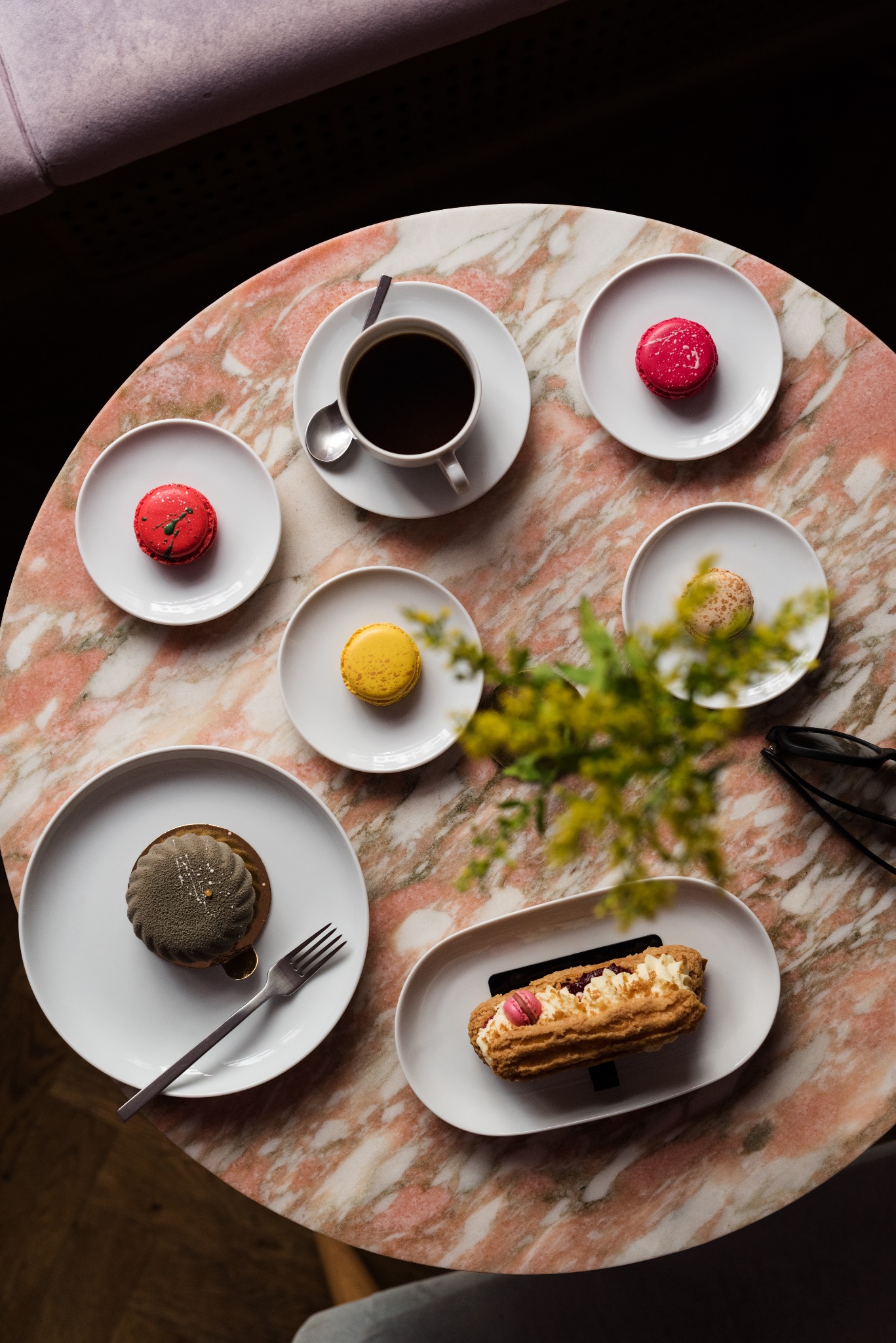 Widok z góry na marmurowy stolik z filiżanką kawy i talerzykami z różnokolorowymi makaronikami, ciastkiem z kremem i ciemnym deserem z widelcem, uzupełniony drobnymi żółtymi kwiatami i okularami.