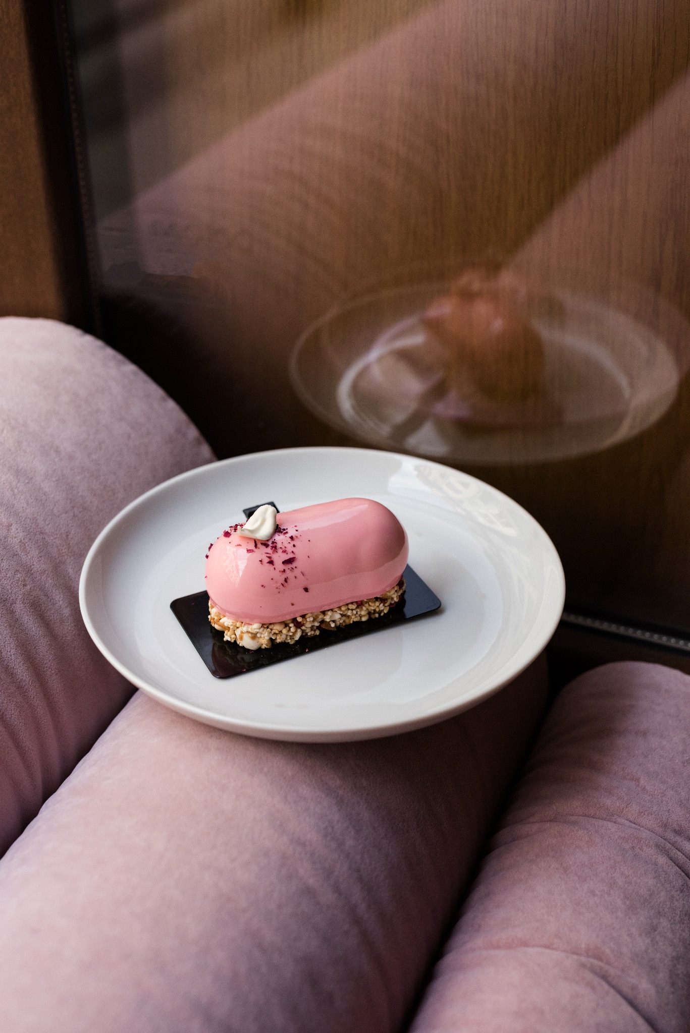 Różowe ciastko w kształcie pastylki z lustrzaną glazurą, posypane okruchami, na białym talerzu, leżącym na różowej sofie, z widocznym odbiciem w oknie.
