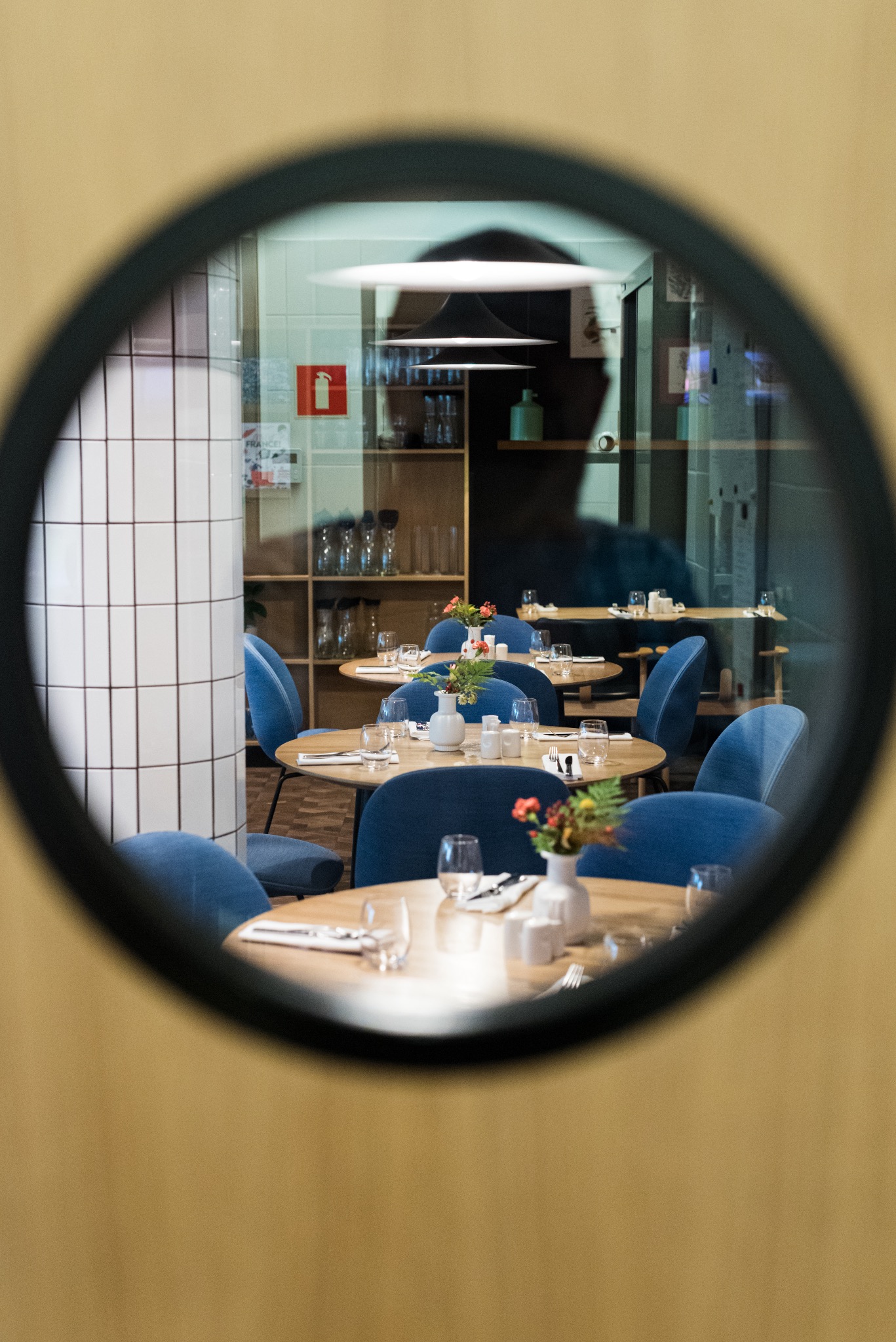 Wnętrze restauracji widziane przez okrągłe okno: okrągłe stoliki z niebieskimi krzesłami, nakryte do posiłku, z małymi wazonami z kwiatami.