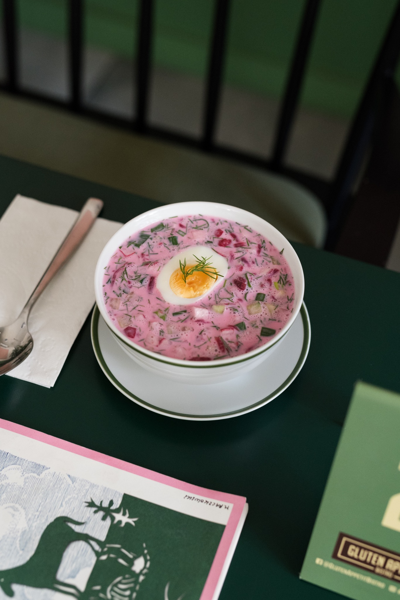 Chłodnik litewski w białej misce na spodku, udekorowany koperkiem i połówką jajka na twardo, obok srebrna łyżka i menu na zielonym stole.