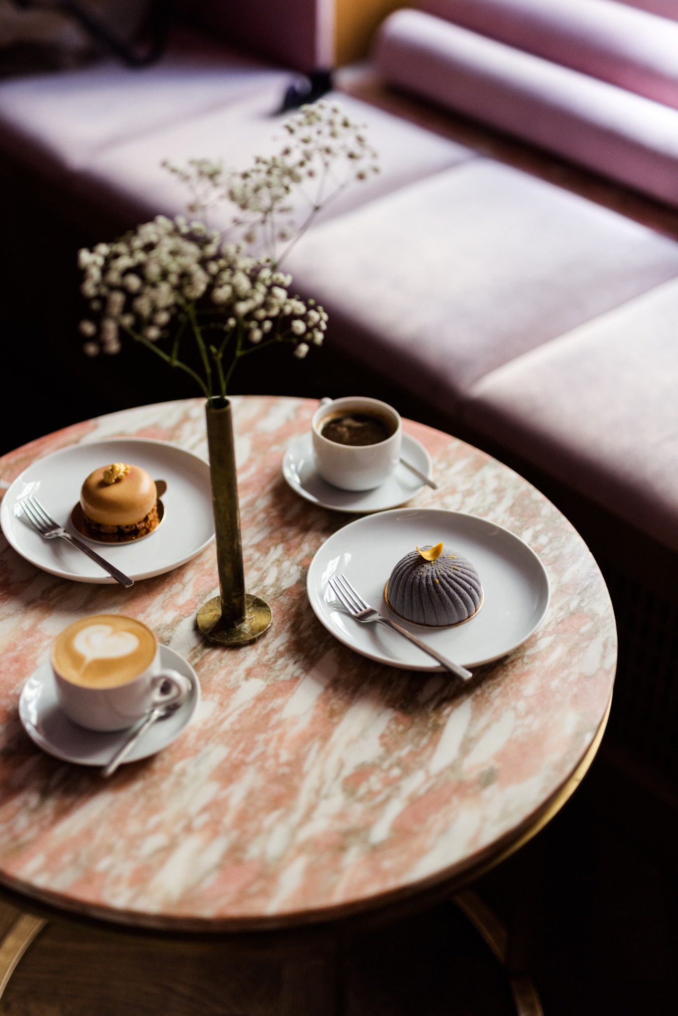 Dwa eleganckie desery na białych talerzach, latte z sercem i czarna kawa na marmurowym stoliku w kawiarni, dekorowane gałązką gipsówki w metalowym wazonie.