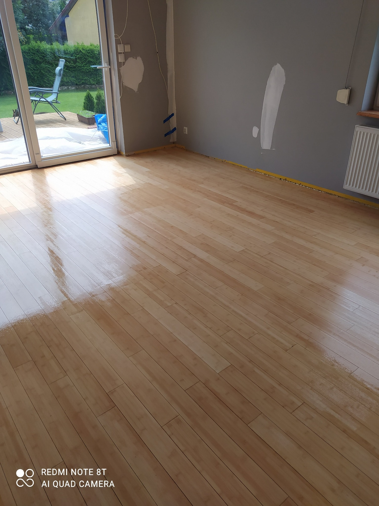 Świeżo polakierowany, błyszczący parkiet w jasnym odcieniu drewna, widoczne ślady po taśmie malarskiej przy ścianach, przygotowane do malowania ściany w szarym kolorze, widok na ogród przez otwarte...