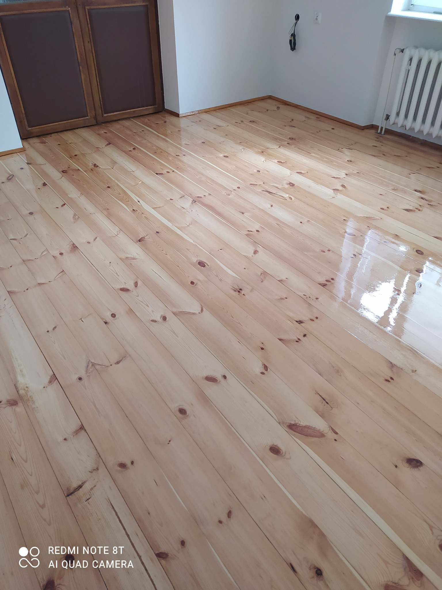 Lakierowana podłoga z jasnego drewna ułożona w deski, widoczne drzwi w tle i fragment grzejnika