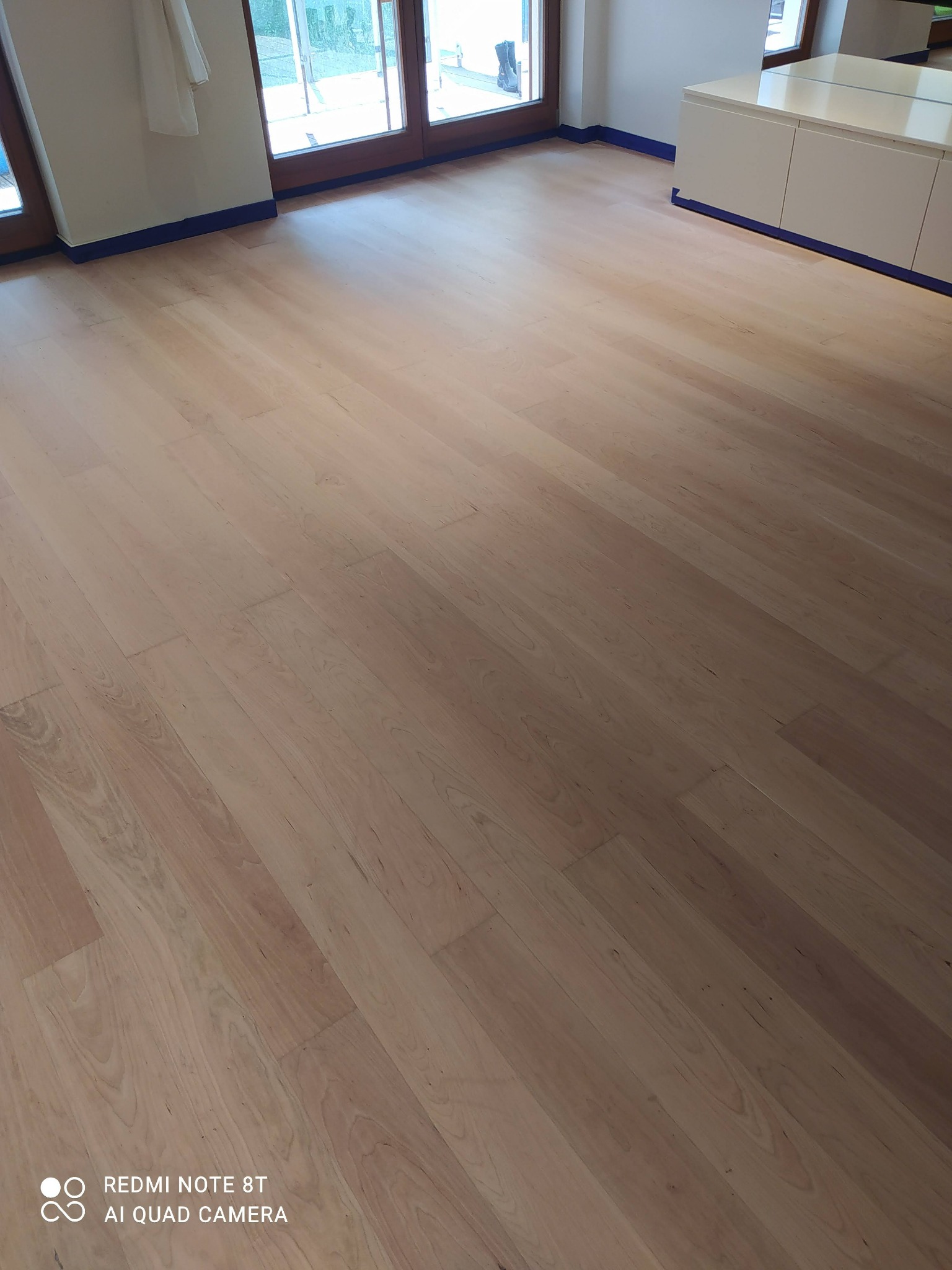 Jasny, świeżo ułożony parkiet w pomieszczeniu z listwami przypodłogowymi zabezpieczonymi niebieską taśmą malarską, widoczne drzwi balkonowe i biała komoda.