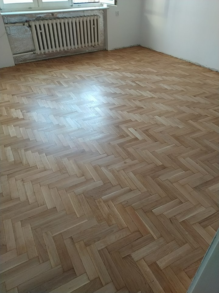 Ułożony parkiet w jodełkę w jasnym odcieniu drewna, widoczny grzejnik i fragment okna w tle, surowe ściany wymagające wykończenia.