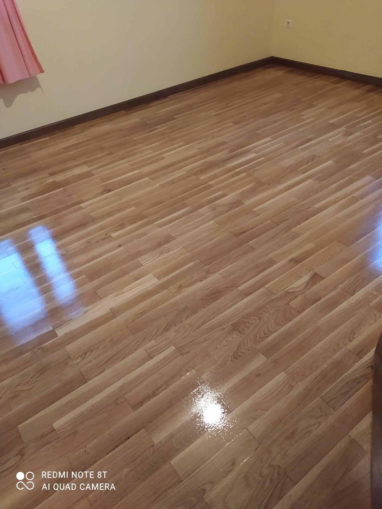 Ułożone panele podłogowe imitujące drewno w jasnym odcieniu, widoczny fragment ściany z brązową listwą przypodłogową i różowy fragment zasłony w rogu.