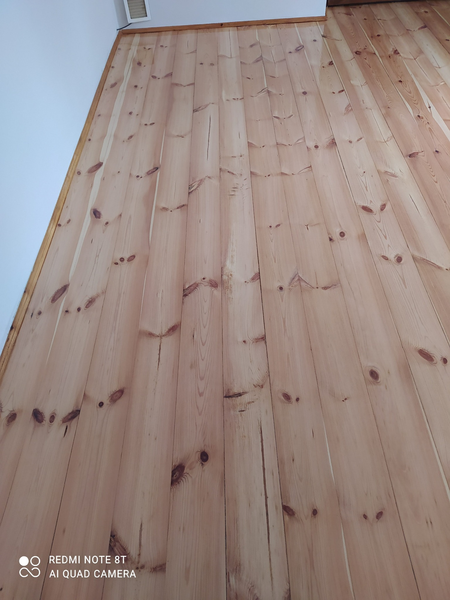Ściana wykończona jasnymi, pionowo ułożonymi panelami drewnianymi z widocznymi sękami, sfotografowana pod kątem od dołu, z fragmentem białego sufitu widocznym w lewym górnym rogu.