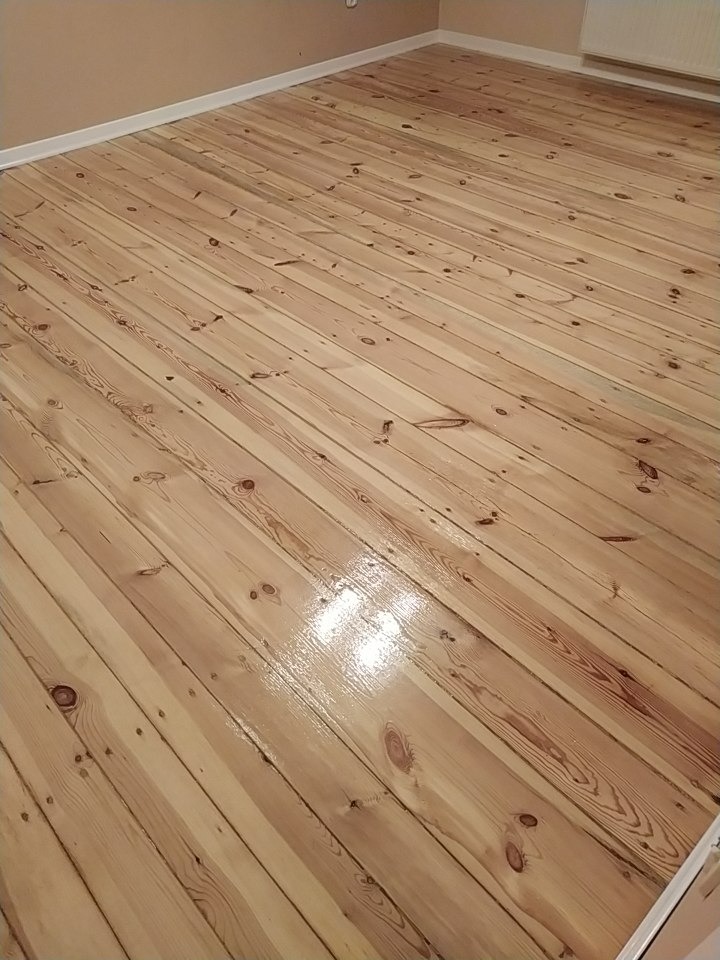 Świeżo polakierowana, jasna drewniana podłoga z widocznymi słojami, ułożona ukośnie w pomieszczeniu z beżowymi ścianami i białą listwą przypodłogową.