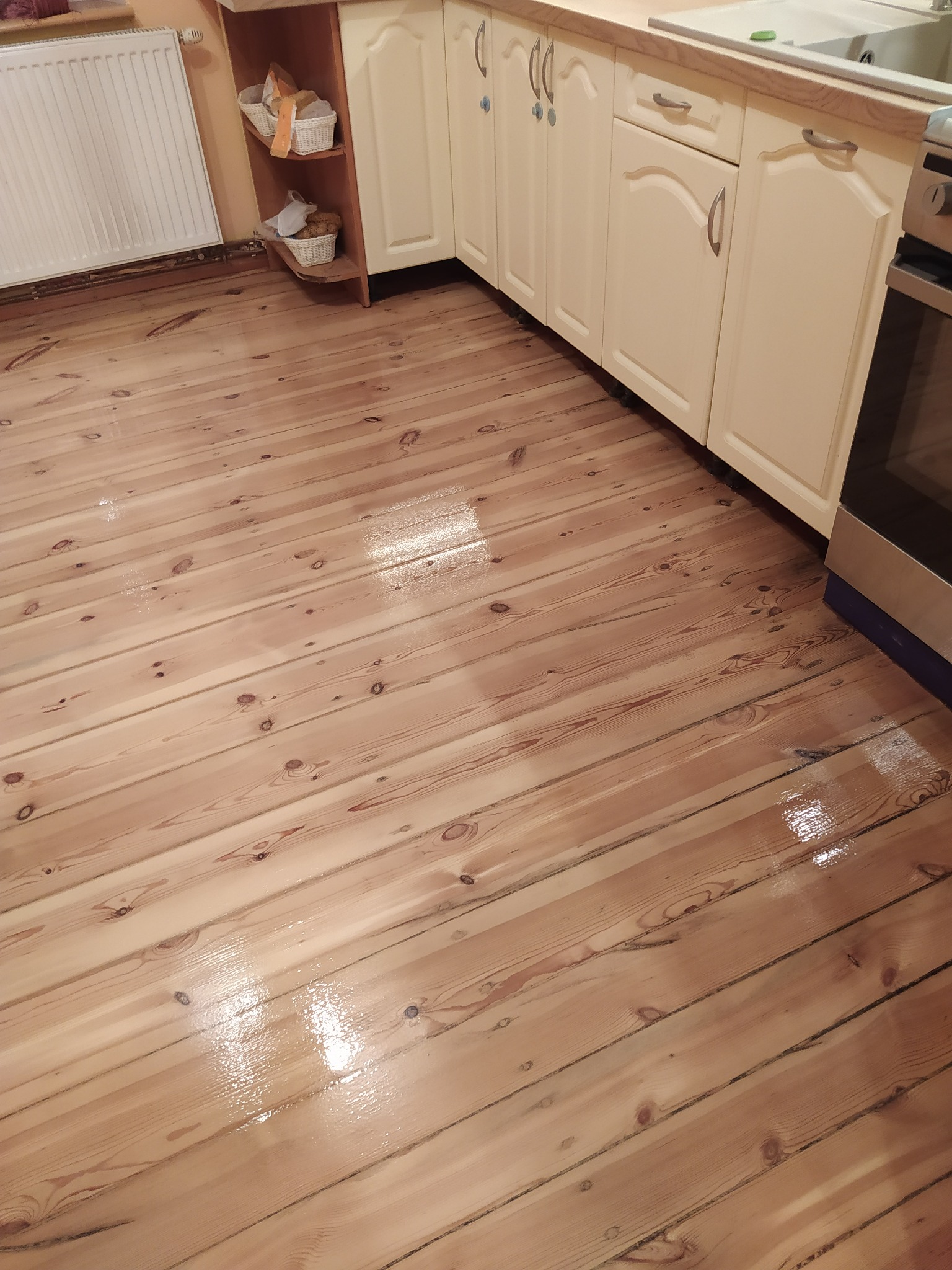 Świeżo polakierowana podłoga z desek w kuchni, widoczne odbicia światła na lakierowanej powierzchni, jasne drewno, kremowe szafki kuchenne w tle.