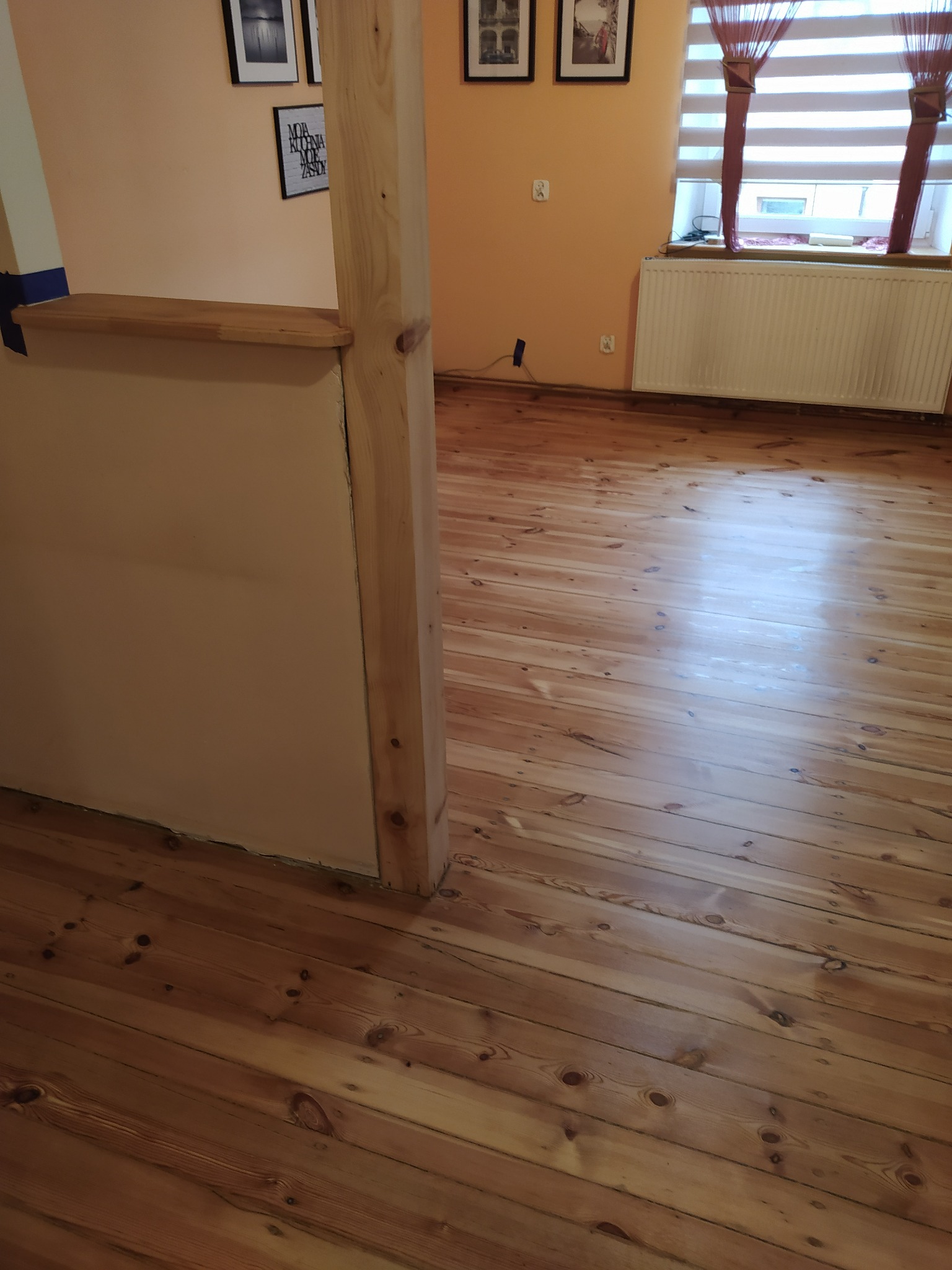 Odnowiona, lakierowana podłoga z desek w pomieszczeniu z jasnymi ścianami i grzejnikiem pod oknem.