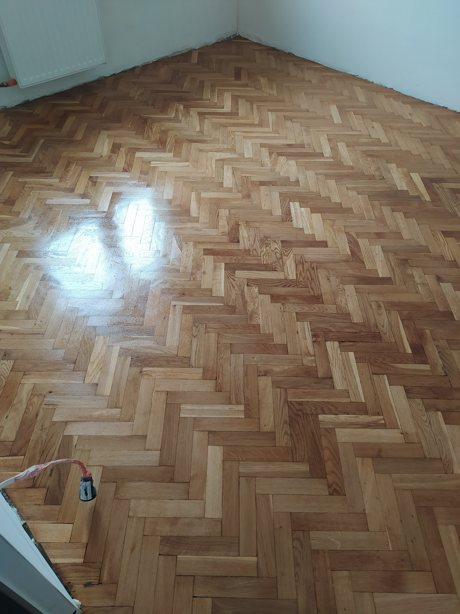 Ułożony parkiet w jodełkę z widocznymi refleksami światła, przy ścianie widoczny kabel z wtyczką.