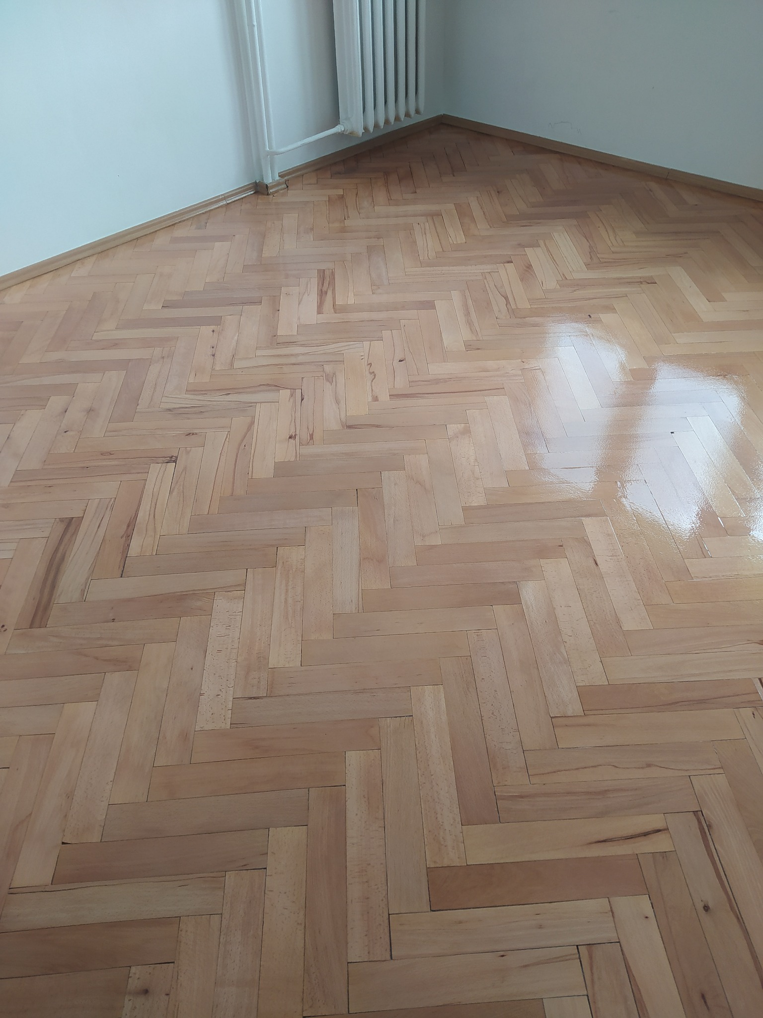 Świeżo polakierowany parkiet ułożony w jodełkę, widoczny fragment ściany z grzejnikiem.