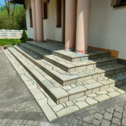 Kamienne schody wejściowe do domu z kolumnami, wykonane z nieregularnych płyt w odcieniach beżu i szarości, prowadzące z kostki brukowej. Widoczne niedokończone wykończenie elewacji obok schodów.