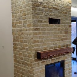 SODZIK-SODOWANIE - Kominek obłożony piaskowcem z drewnianą półką, widoczne otwory wentylacyjne. Jasne, naturalne kolory kamienia i drewna. Częściowo widoczna osoba w tle.