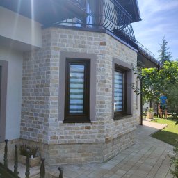 SODZIK-SODOWANIE - Elewacja domu wykończona jasną, nieregularną kostką. Widoczne dwa okna z żaluzjami poziomymi, fragment balkonu z ozdobną balustradą i chodnik z kostki brukowej.