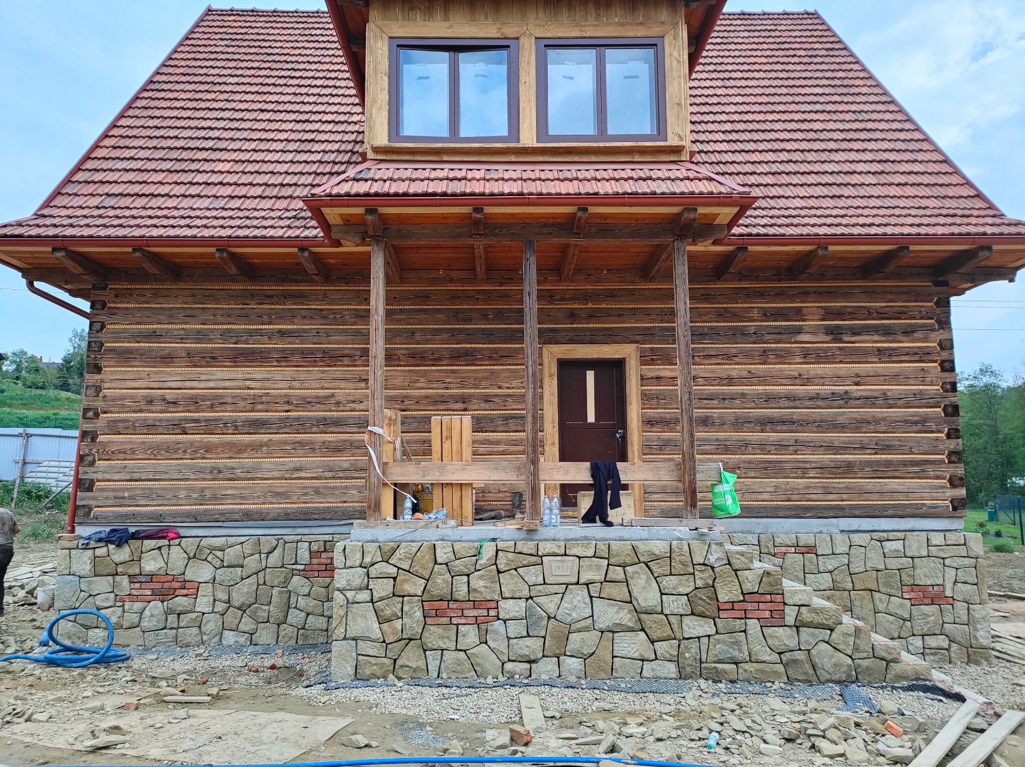 Dom z bali z kamienną podmurówką w trakcie budowy. Widoczne elementy drewniane, dach z dachówki i kamienna elewacja. Na pierwszym planie gruz i materiały budowlane.