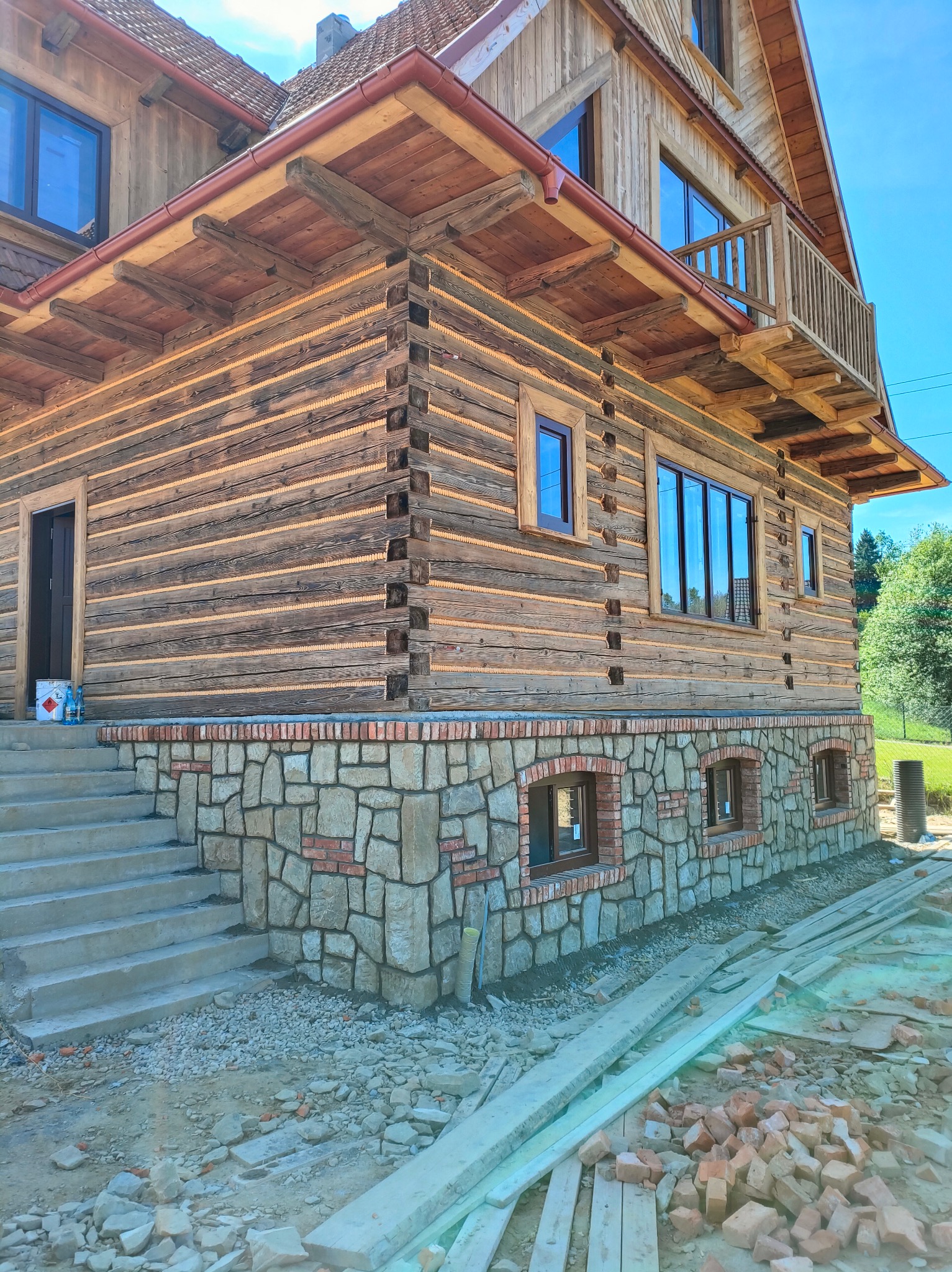 Drewniany dom z kamienną podmurówką i betonowymi schodami w trakcie budowy. Widoczne materiały budowlane i narzędzia. Jasne niebo w tle.