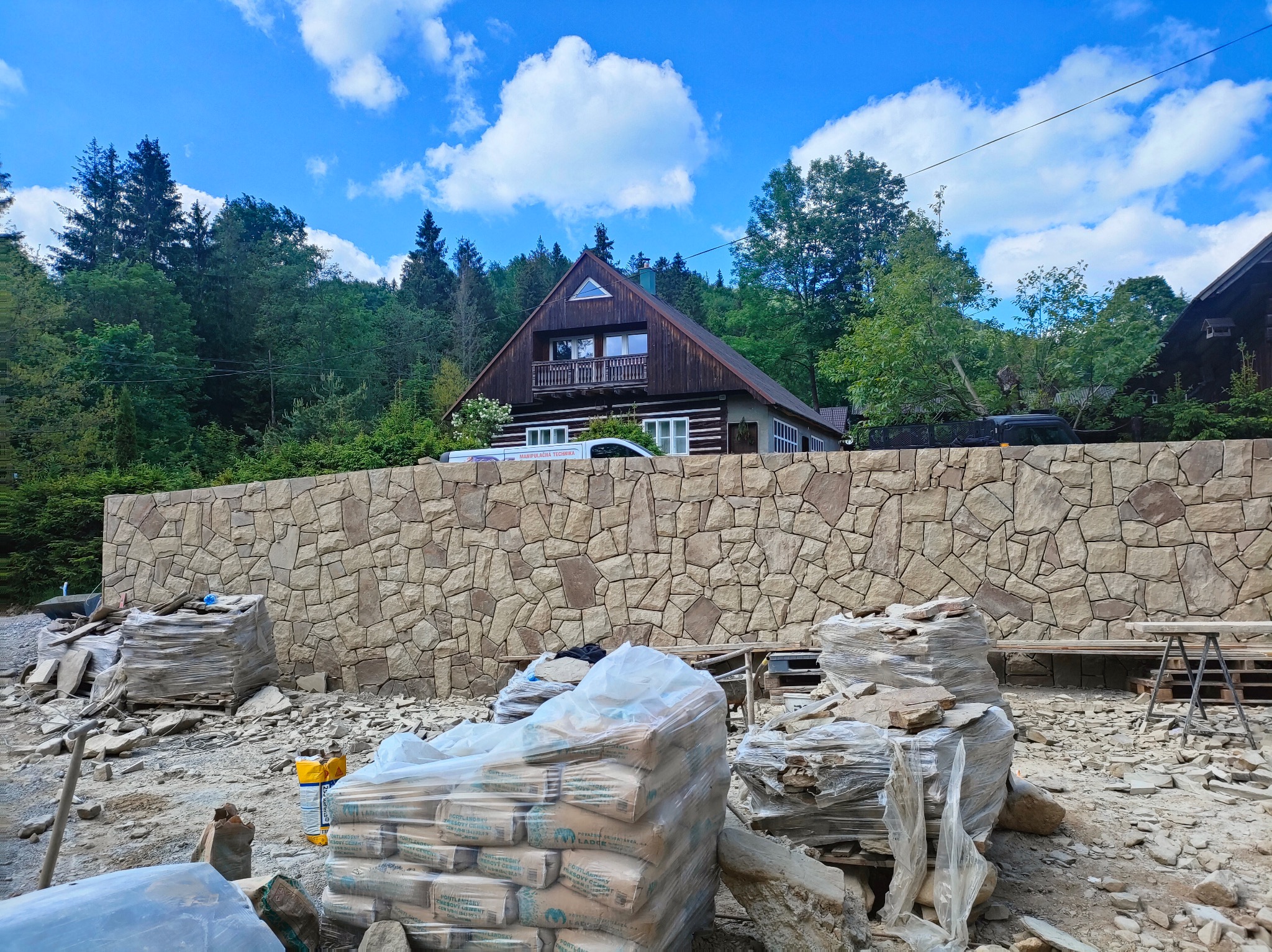 Budowa kamiennego muru oporowego przed domem. Widoczne worki z cementem i materiały budowlane. W tle dom z drewnianą elewacją i zielone drzewa.