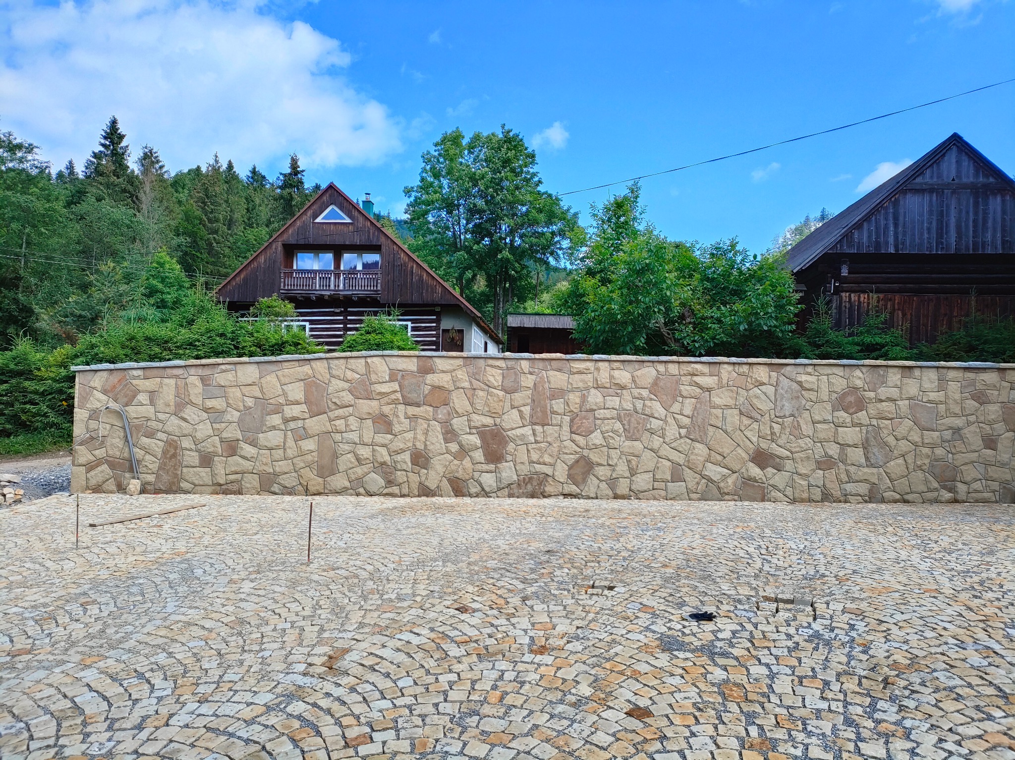 Ułożona kostka granitowa w łukowaty wzór, z kamiennym murem w tle i fragmentami budynków. Widoczne pręty i narzędzia sugerują trwające prace.