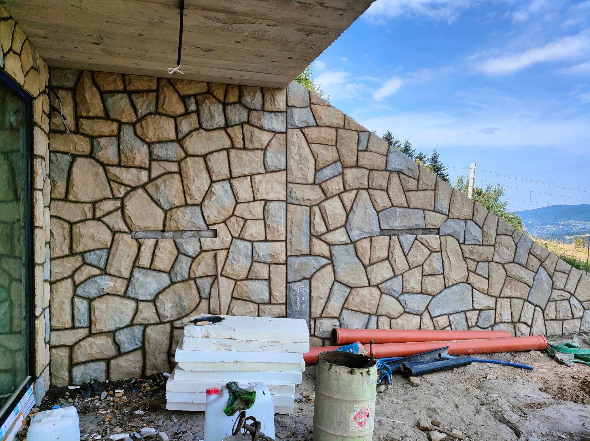 Ściana budynku wykończona kamieniem naturalnym o nieregularnym kształcie, widoczne materiały budowlane i fragment krajobrazu w tle. Detal wykończenia elewacji.