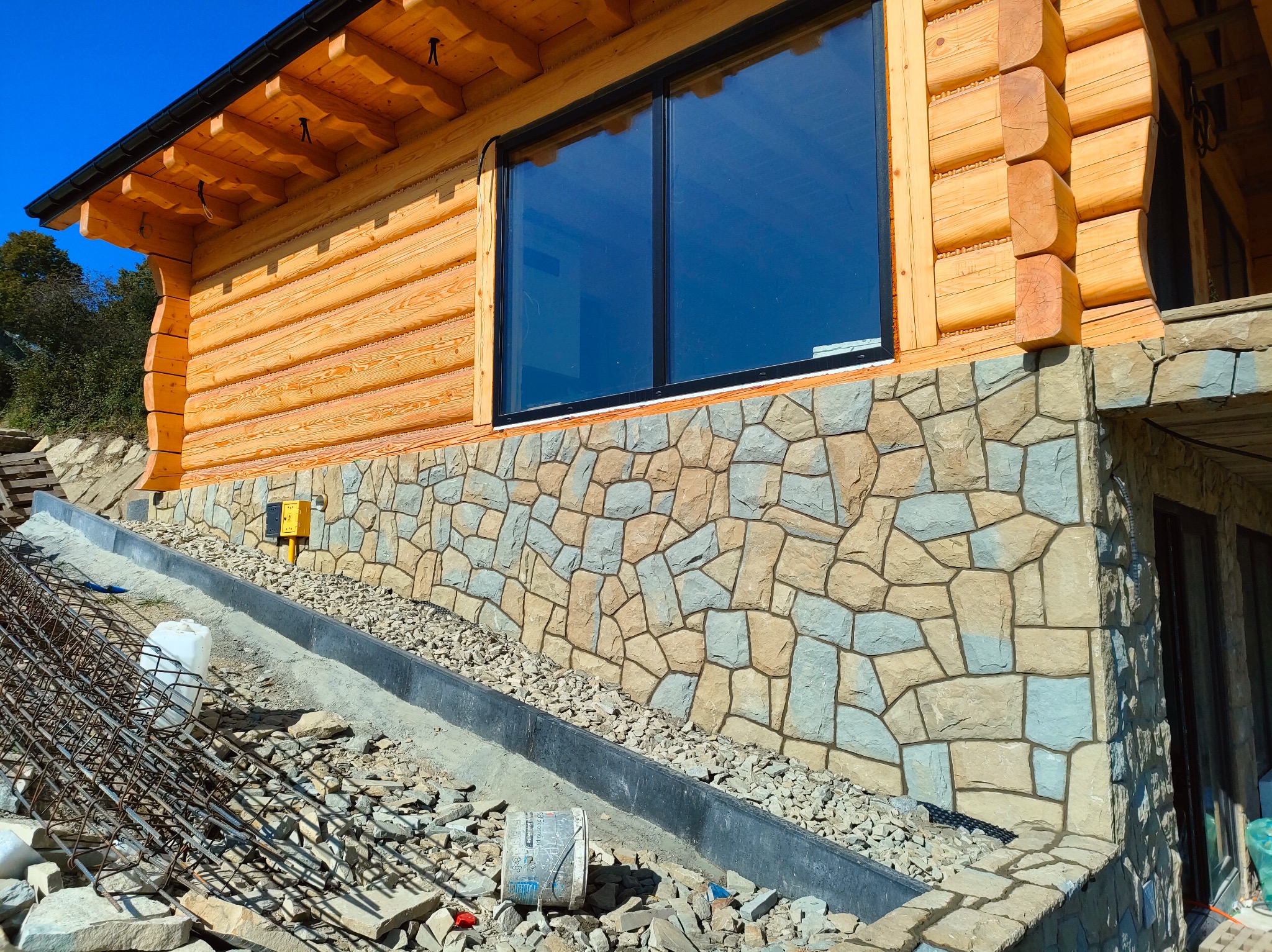 Dom z bali z kamienną elewacją w trakcie budowy. Widoczne zbrojenie, gruz i materiały budowlane. Duże okno odbija niebo. Elementy drewniane w ciepłym odcieniu.