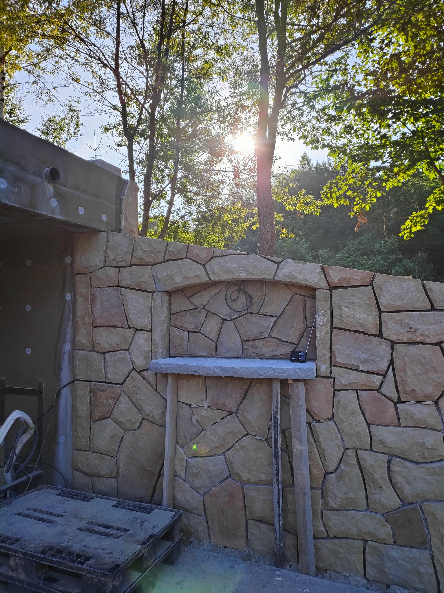 Kamienna ściana w ogrodzie, z elementami dekoracyjnymi i półką. Widoczne drzewa i słońce w tle. Częściowo otynkowana ściana z lewej strony. Paleta budowlana na dole.