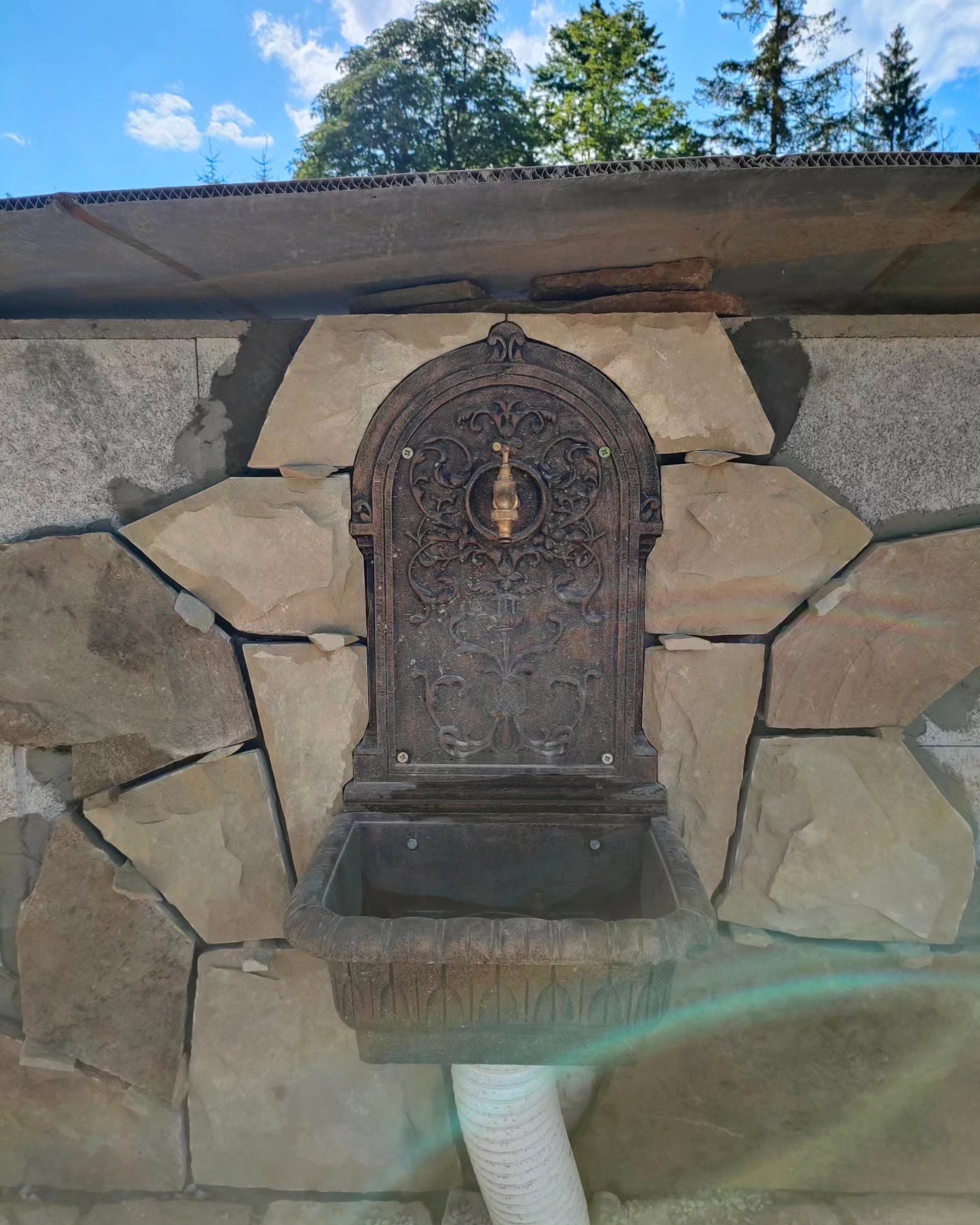 Ozdobna, kamienna fontanna ogrodowa z mosiężnym kranem, wkomponowana w mur z nieregularnych kamieni. Realizacja w Lubieniu.