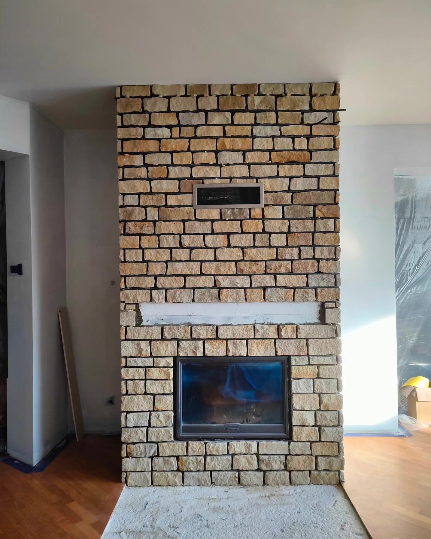 Kamienny kominek w trakcie budowy, z widocznym wkładem kominkowym i fragmentami niezakończonej obudowy. Surowe wykończenie, ciepłe odcienie kamienia.