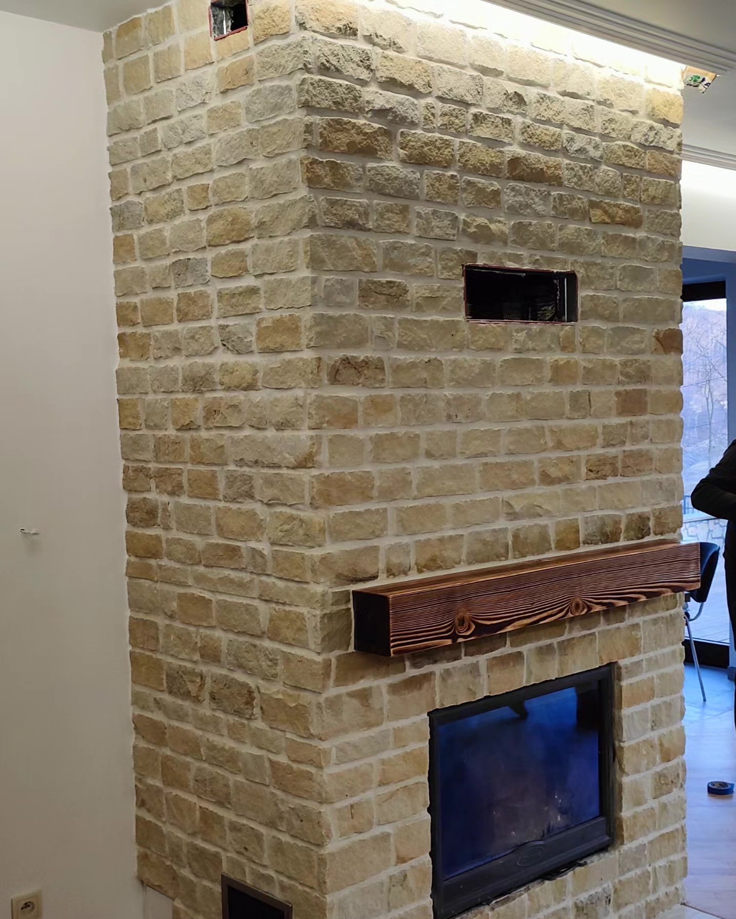 Kominek obłożony piaskowcem z drewnianą półką, widoczne otwory wentylacyjne. Jasne, naturalne kolory kamienia i drewna. Częściowo widoczna osoba w tle.