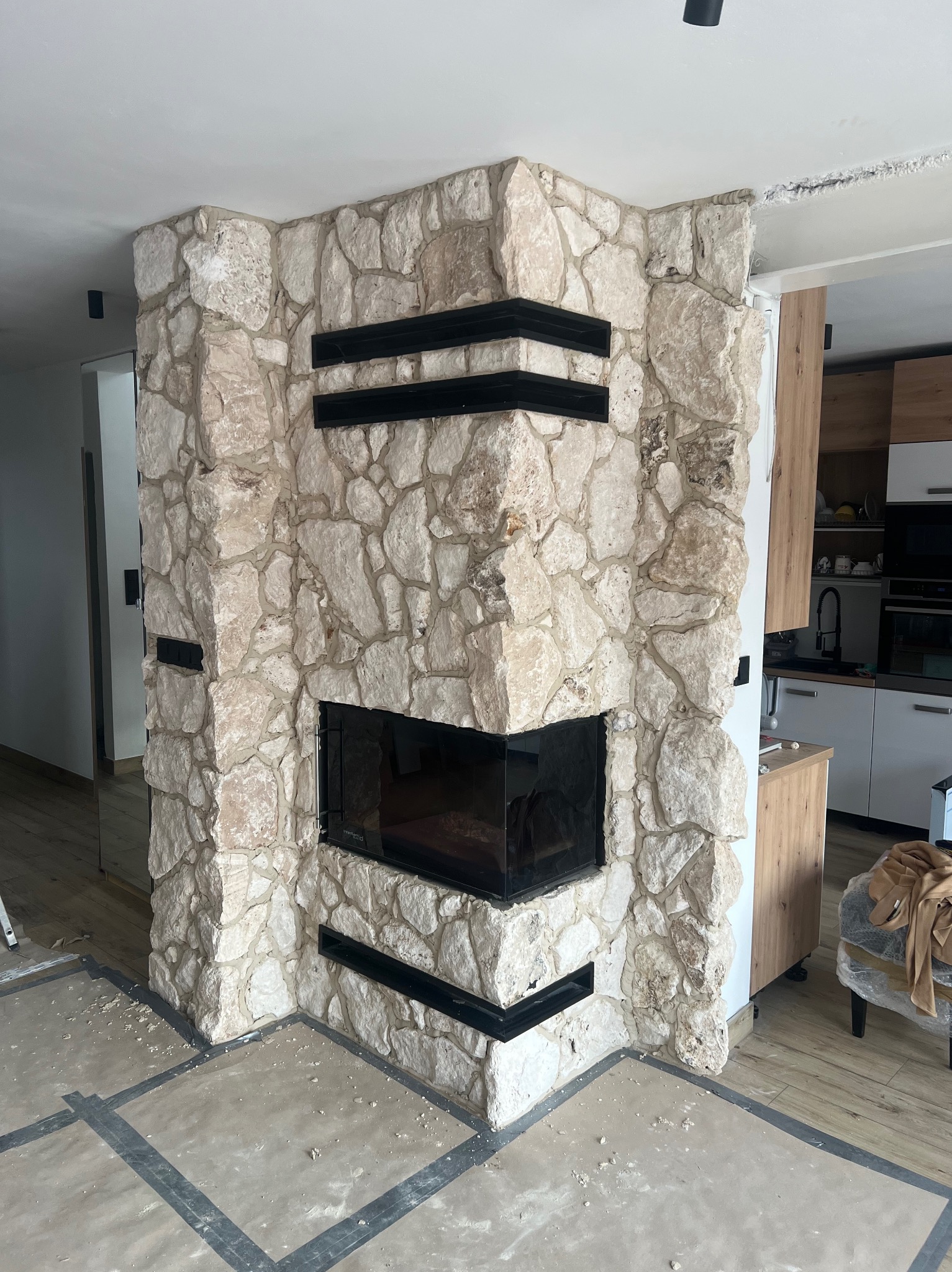 Kamienny kominek narożny z czarnymi, poziomymi akcentami, w trakcie wykończenia. Widoczne fragmenty podłogi z szarymi liniami i elementy kuchni w tle.