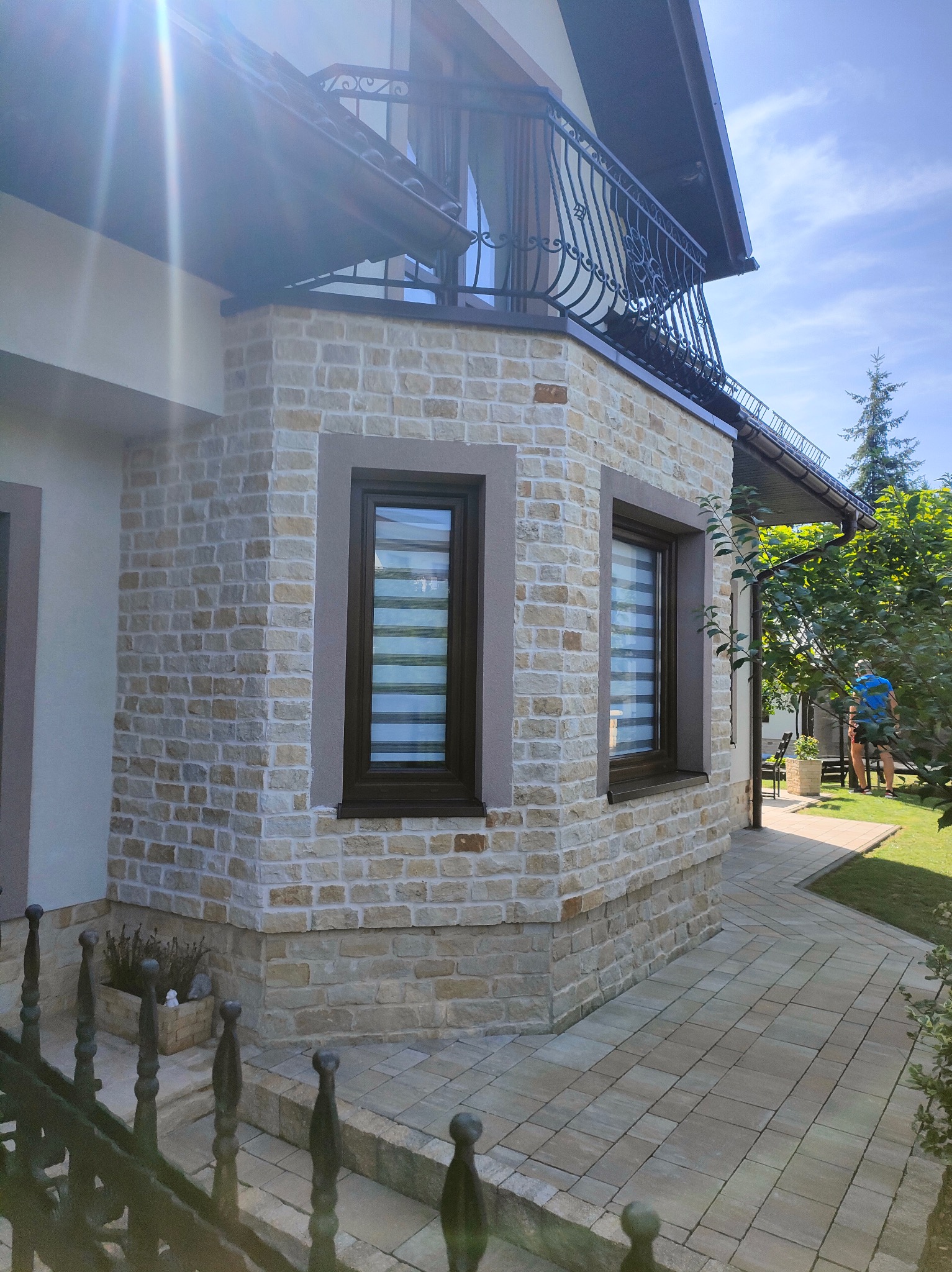 Elewacja domu wykończona jasną, nieregularną kostką. Widoczne dwa okna z żaluzjami poziomymi, fragment balkonu z ozdobną balustradą i chodnik z kostki brukowej.