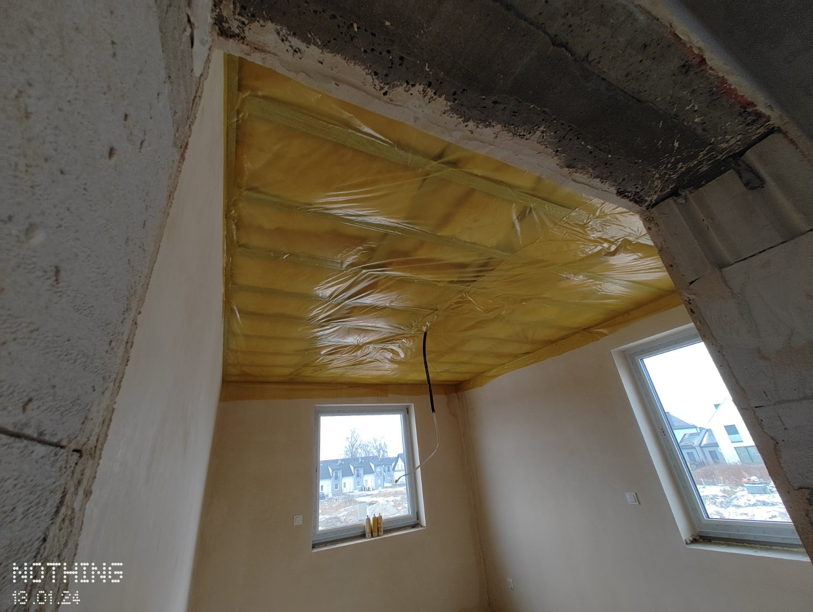 Montaż izolacji z folią na suficie podwieszanym w pomieszczeniu z oknami, widoczne surowe ściany i wystające przewody elektryczne.