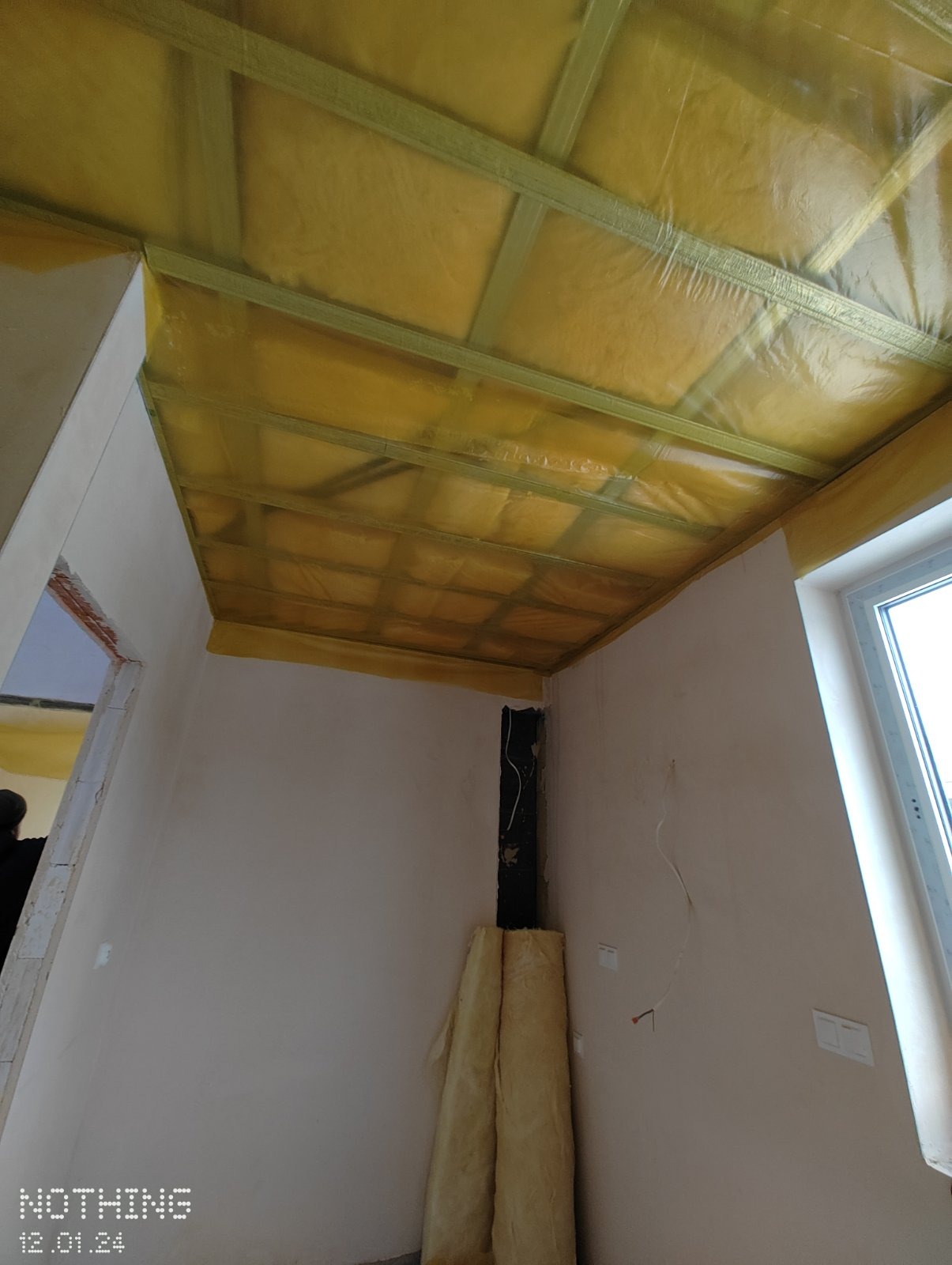 Montaż izolacji termicznej z folią na suficie podwieszanym w trakcie prac wykończeniowych, widoczne profile konstrukcyjne i rolki materiału izolacyjnego przy ścianie.