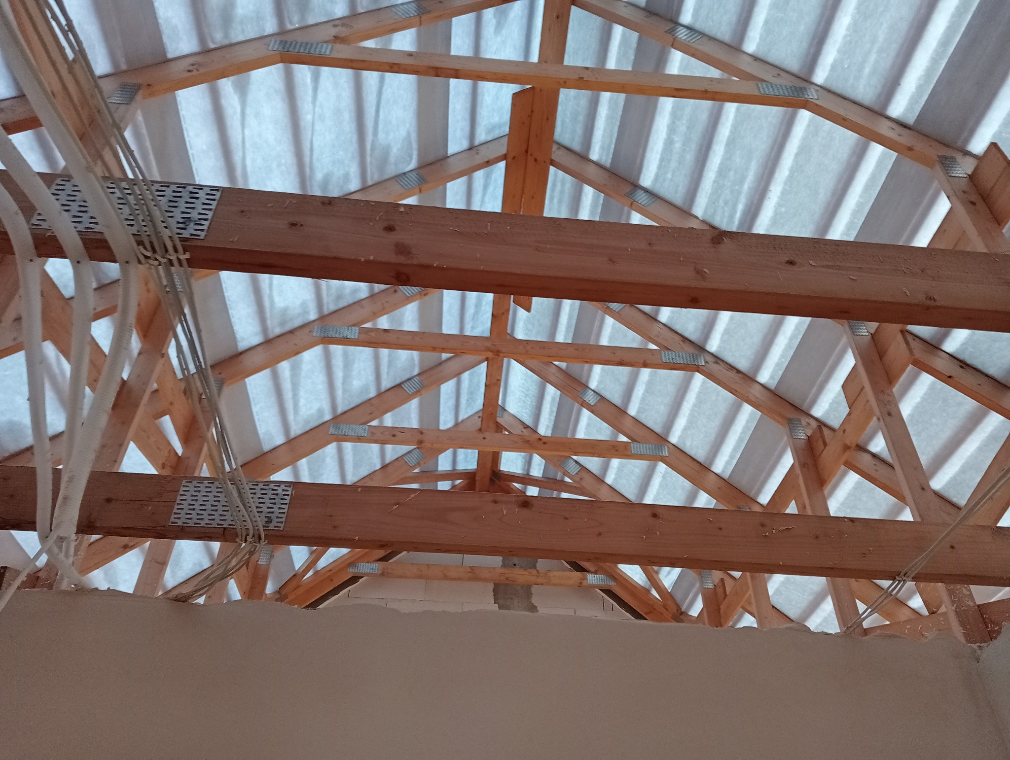 Konstrukcja drewniana dachu z widocznymi elementami mocującymi i przewodami elektrycznymi, widok od wewnątrz budynku w trakcie budowy.