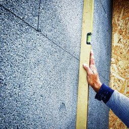 Renovieren Andrzej Nawrocki - Ręka przytrzymuje poziomicę na szarej, chropowatej powierzchni ściany, obok widoczna płyta OSB.