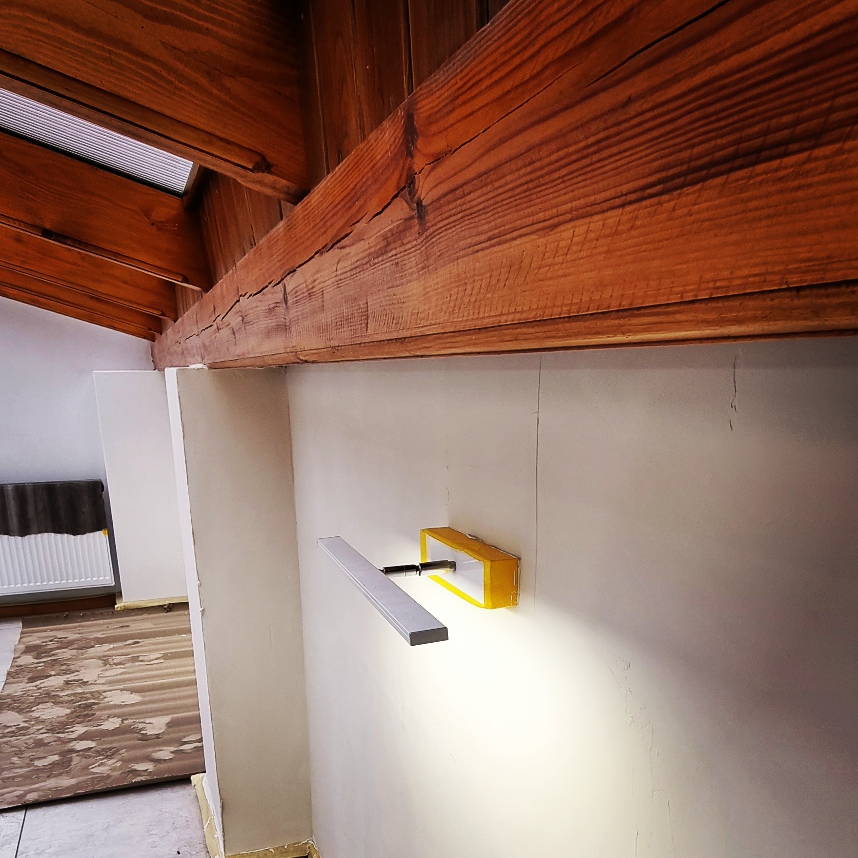 Przygotowanie do montażu lampy na ścianie w pomieszczeniu z widoczną drewnianą konstrukcją stropu; na ścianie widoczne ślady po wcześniejszych pracach.